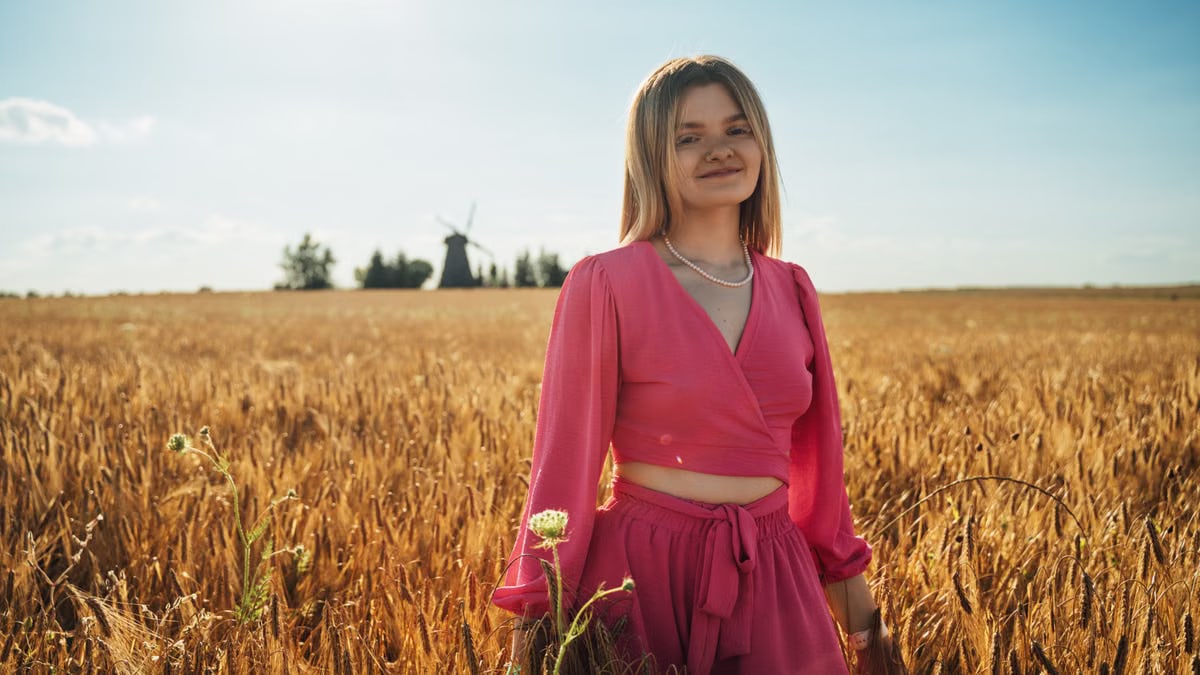 A young woman in pink stands joyfully in a golden wheat field at sunset.